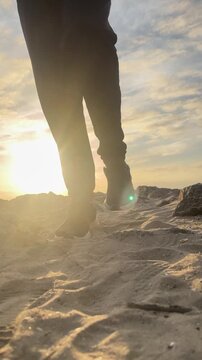 A person is walking on a beach with their feet in the sand. The sky is cloudy and the sun is setting