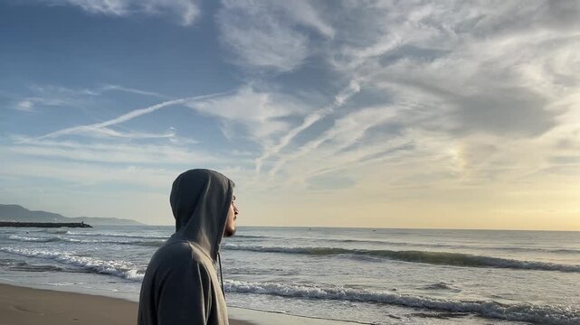 A man is standing on the beach with his hood up. The sky is cloudy and the ocean is calm