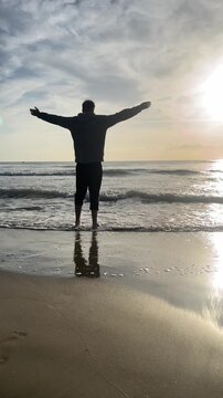 A man stands on the beach looking out at the ocean. The sky is cloudy and the sun is setting