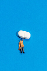 Miniature figure struggles to lift oversized pill on bright blue background