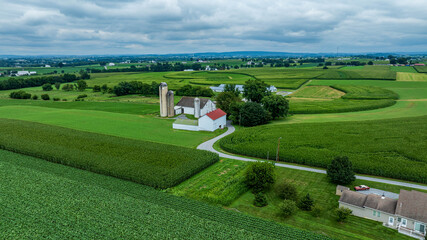 A beautiful view of expansive farmland featuring a white barn and silos surrounded by lush green fields. Rolling hills and a cloudy sky create a serene atmosphere in the countryside. © Greg Kelton