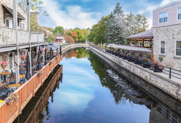 Two restaurant patios on opposite sides of a small urban canal, restaurant seating, stone buildings, autumn foliage, calm water reflecting clouds, daytime, nobody