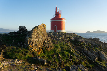 Punta Robaleira red lighthouse in Home cape at sunset in Rias Baixas zone in Galicia coast with Cíes islands in the background.