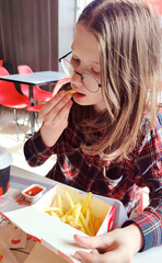 Young girl in glasses sits in a cafe and eats French fries. Delicious unhealthy food.