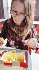 Young girl in glasses sits in a cafe and eats French fries. Delicious unhealthy food.