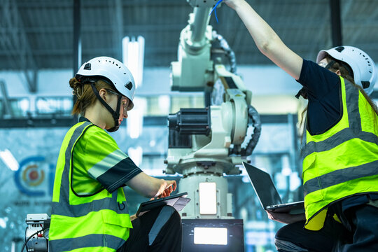 Robotics technicians troubleshoot a faulty industrial arm. One engineer runs diagnostics on a laptop while the other uses a teach pendant to inspect the robot's wiring and servo motors.