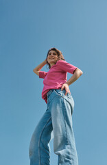 Beautiful carefree young woman with curly hair in pink t-shirt smiling and looking down at camera on blue sky background low angle shot.
