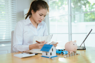 Stressed businesswoman sitting at desk, worrying about financial problems, calculating expenses with piggy bank, coins, and house model on table.