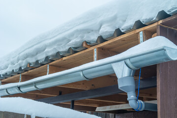 The roof, covered with metal profiles, is covered with snow in winter. The water drainage system from the roof of the house is covered with snow. А rural country house in the winter cold.