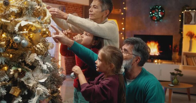 Cheerful grandparents and two little granddaughters decorating Christmas tree together. Joyful multigenerational family bonding during festive holiday season at home.