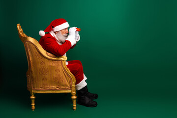 Smiling Santa Claus in a red suit sitting on a golden chair and peering through toy binoculars against a green background