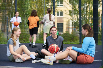 Several happy restful teenage girls sitting on sports court talking and relaxing after streetball game against their friends playing basketball