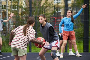 Group of teenage Caucasian girls playing streetball on outdoor court, one girl dribbling basketball while another girl defending, teammates standing in background watching action