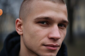 Close-up of a young man with a shaved head, wearing a dark hoodie, looking directly at the camera with a serious expression and soft focus background