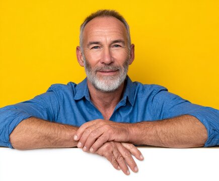 A mature man with a gray beard and a blue shirt, leant against a white backdrop against a vivid yellow background, beams with a friendly smile.