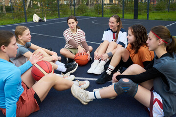 Group of six teenage girls sitting in circle on outdoor basketball court holding basketballs, smiling and talking after streetball game, diverse hairstyles and athletic wear visible