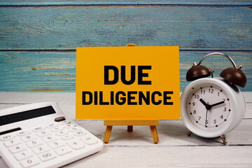 Due Diligence text written on paper card with calculator and alarm clock on wooden background