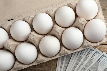 Raw chicken eggs in egg carton and dollar banknotes on wooden table, flat lay