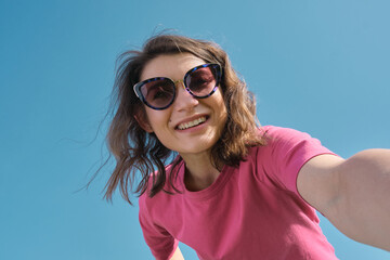 Beautiful carefree young woman in sunglasses and pink t-shirt taking selfie on camera on blue sky background low angle shot.