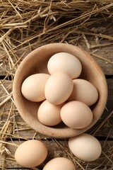 Raw chicken eggs in bowl and straw on wooden table, closeup