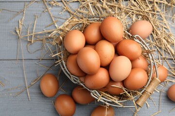 Raw chicken eggs, metal basket and straw on grey wooden table, flat lay