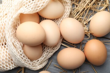 Raw chicken eggs, mesh bag and straw on grey wooden table, flat lay
