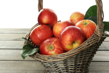 Fresh apples in wicker basket and green leaves on wooden table against white background