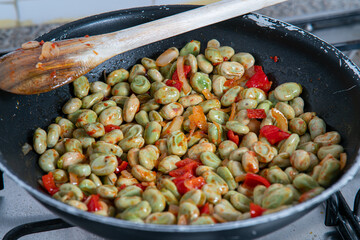 Sautéed broad beans with red bell pepper cooking in a frying pan on a gas stove — a traditional Mediterranean spring dish rich in color and texture.
