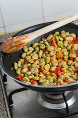 Sautéed broad beans with red bell pepper cooking in a frying pan on a gas stove — a traditional Mediterranean spring dish rich in color and texture.