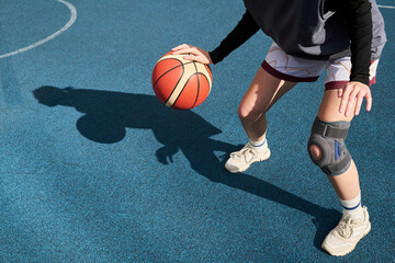 Teenager Caucasian girl dribbling basketball on outdoor court wearing knee brace, focusing on ball control during streetball game, lower body and hands visible in action