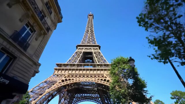 An extreme low angle video of the Eiffel Tower's wrought iron structure ascending towards a cerulean sky, framed by Haussmannian buildings and lush trees in crisp midday sunlight. Serene and grand