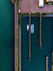 Aerial view of marina two boats docked between parallel wooden piers in green blue water, bordered by rocky shore, red roofed building, and colorful structures in a compact harbor layout.