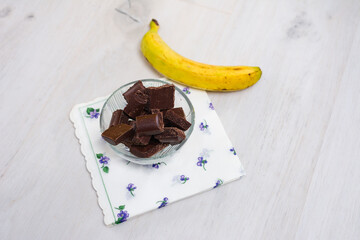 Chocolate chunks in a glass bowl with a banana on a floral napkin.