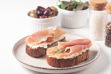 Two bruschettas with prosciutto, ricotta cheese and rucola on ciabatta bread. Olive and spices  decorated.