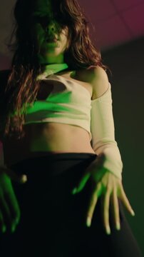 A female dancer with long brown hair, wearing a white top and black shorts, performs a sensual dance in a dimly lit studio. Sensual Dance Performance. Young Woman Dancing Sensually in Neon Lit Studio