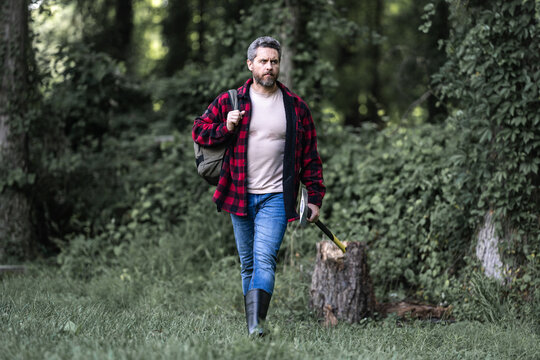 The Lumberjack holds axe with serious face on the forest. A lumberjack with a large ax examines the tree before felling. Serious lumberjack. Confident young bearded man holding a big axe outdoor. - Powered by Adobe