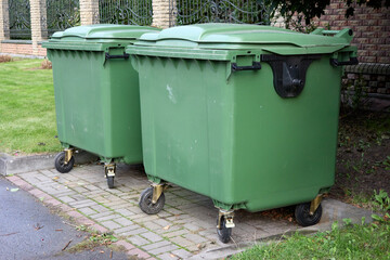 Two green wheelie bins stand ready for trash collection on a paved area near a building, representing municipal waste management and local environmental cleanliness outdoors.