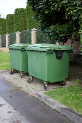 A suburban tableau presents two green industrial trash containers lined up on the curb, juxtaposed with a meticulously maintained green hedge and an elegant wrought iron fence backdrop.