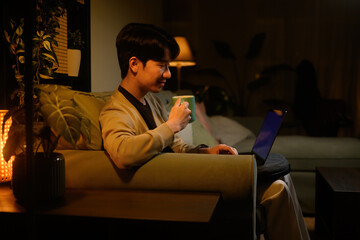 Relaxed remote worker sitting on sofa, smiling while using laptop and drinking coffee in dimly lit living room