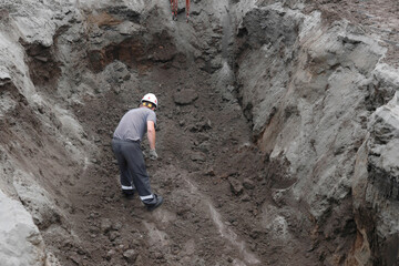 Male worker excavating in deep trench at construction site with helmet and tools.