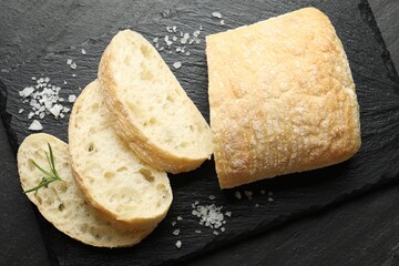 Cut crispy ciabatta, rosemary and salt on black textured table, top view