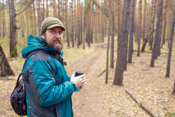A bearded man stands on a path in the forest, turning around, holding a cup of tea in his hand