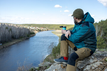 A man sits on a rock overlooking a river, holding a cup of coffee