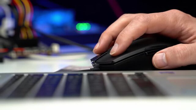 Tight cu video of an adult hand meticulously operating a black computer mouse, clicking scroll wheel and right button, glowing wires and circuit boards in bokeh background. Modern technology concept
