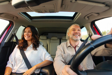 Happy couple sitting in their car, man on drivers seat, spouses going on vacation, looking at road and smiling, view from dashboard