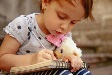 Little girl drawing in her notebook beside a teddy bear.