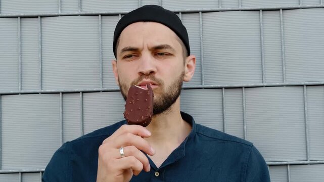 A man in a dark blue shirt and black cap enjoys a chocolate ice cream bar against a metal wall background. Horizontal 4k footage