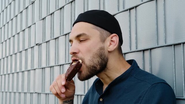 A man in a dark blue shirt and black cap enjoys a chocolate ice cream bar against a metal wall background. Horizontal 4k footage