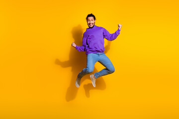 Energetic young man wearing purple hoodie jumps joyfully on vibrant yellow background, expressing...