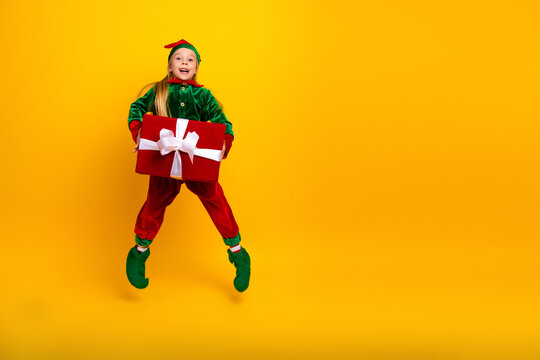 Smiling girl dressed as an elf holding a holiday gift box standing against a yellow background, celebrating Christmas - Powered by Adobe